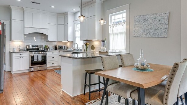 Een moderne keuken met witte kastjes, een centraal eiland, hanglampen en een eethoek met een houten tafel.