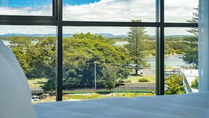 Ein Zimmer mit großem Fenster und Blick auf einen Park, Bäume und ein weiter Wasserblick.