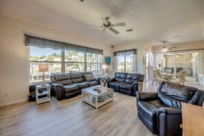 Ein geräumiges Wohnzimmer mit Ledersofas, einem Couchtisch und einem Deckenventilator. Große Fenster bieten Blick auf Palmen.