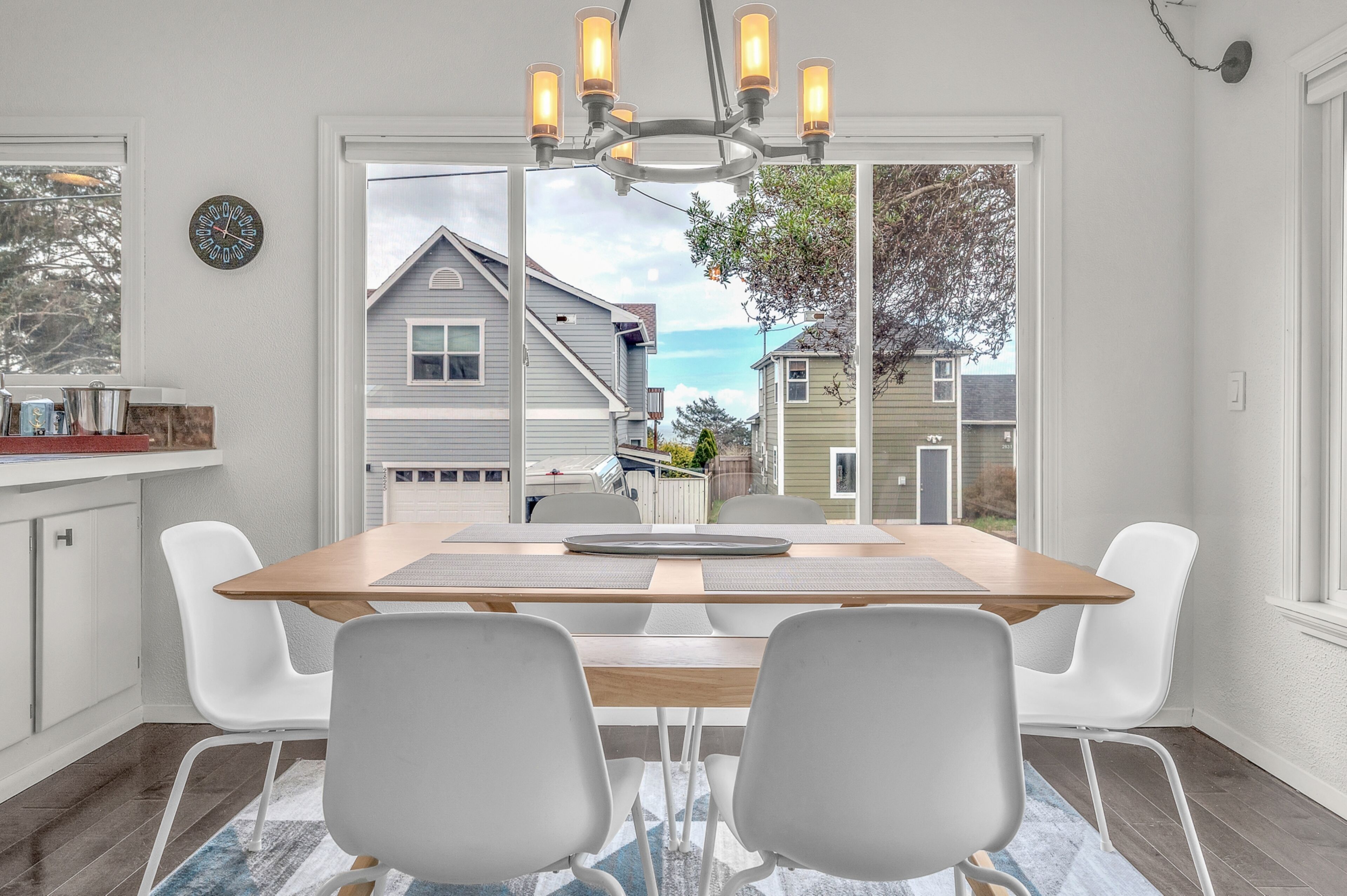 Un coin repas avec une table en bois, des chaises blanches, un lustre et une vue sur les maisons voisines.