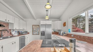 A modern kitchen with white cabinets, a stainless steel refrigerator, a built-in oven, and a large window with a view of trees. - Beach Nest: Modern Coastal Getaway Near Ocean, Trails & Dining (Lincoln City)