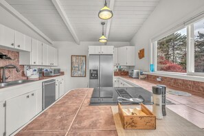 A modern kitchen with white cabinets, a stainless steel refrigerator, a built-in oven, and a large window with a view of trees.