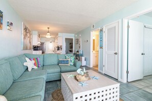 A living room with a teal sofa, a coffee table with decorative items, and a kitchen in the background. - IS256-  Ahhhmazing View, Location X3, Pool, Beach Front, 2nd Floor! (Gulf Shores)