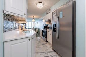 A kitchen with white cabinets, a stainless steel refrigerator, a chandelier, and a tiled floor. - IS256-  Ahhhmazing View, Location X3, Pool, Beach Front, 2nd Floor! (Gulf Shores)