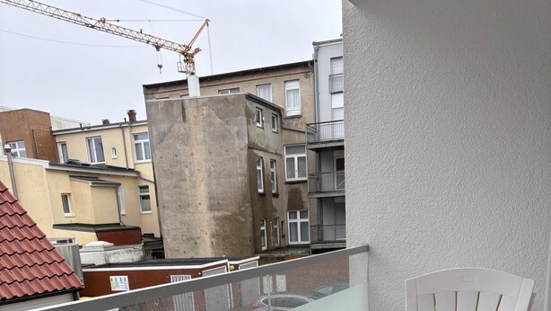 Un balcon avec une chaise blanche, une balustrade en verre et une vue sur des immeubles d’habitation.
