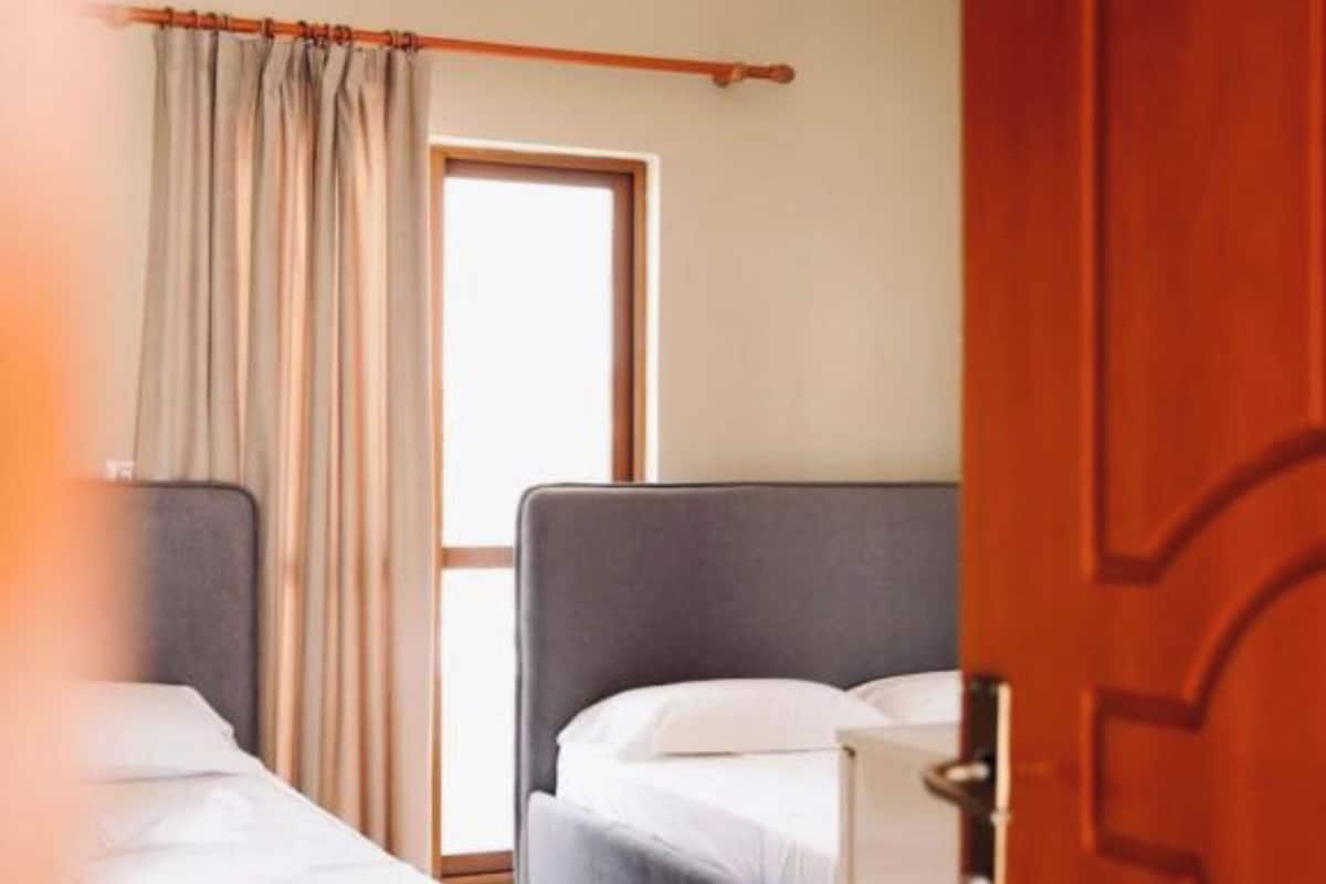 A hotel room with two beds, a wooden door, a window with curtains, and a ceiling light.