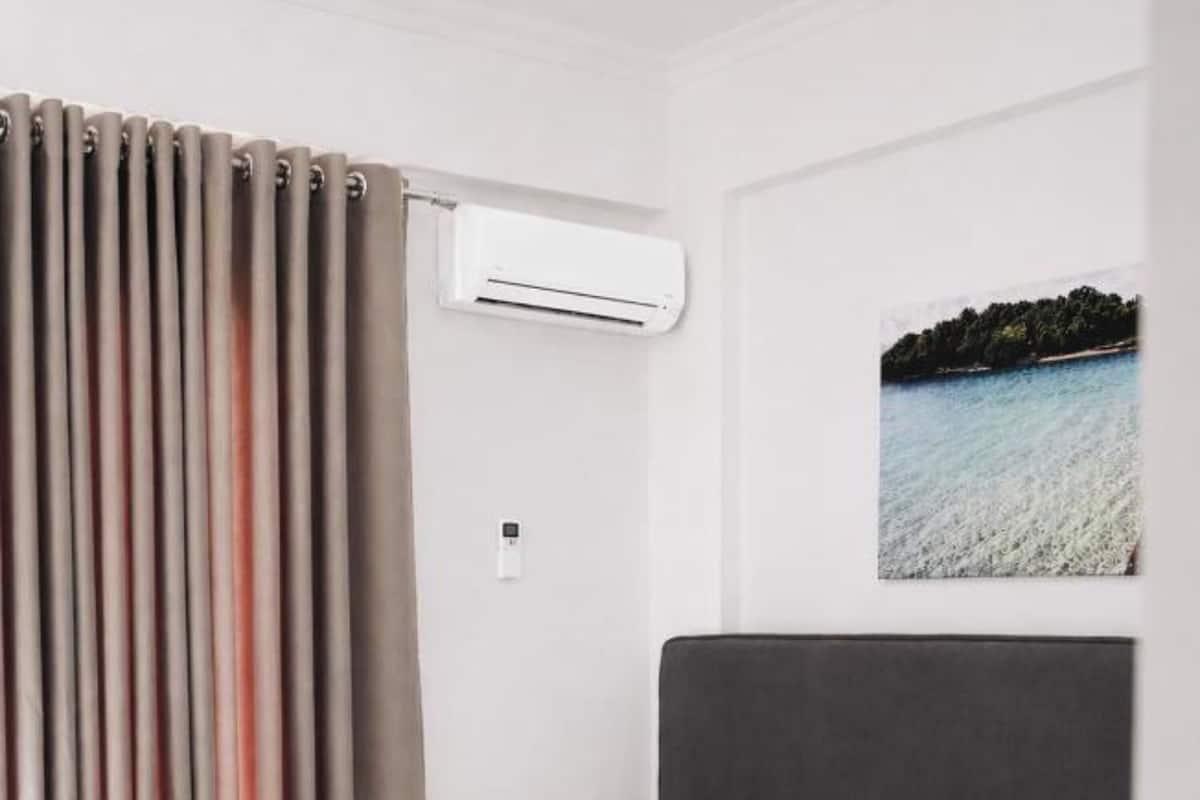 A hotel room with a bed, two bedside tables, an air conditioning unit, and a painting of a coastal landscape.