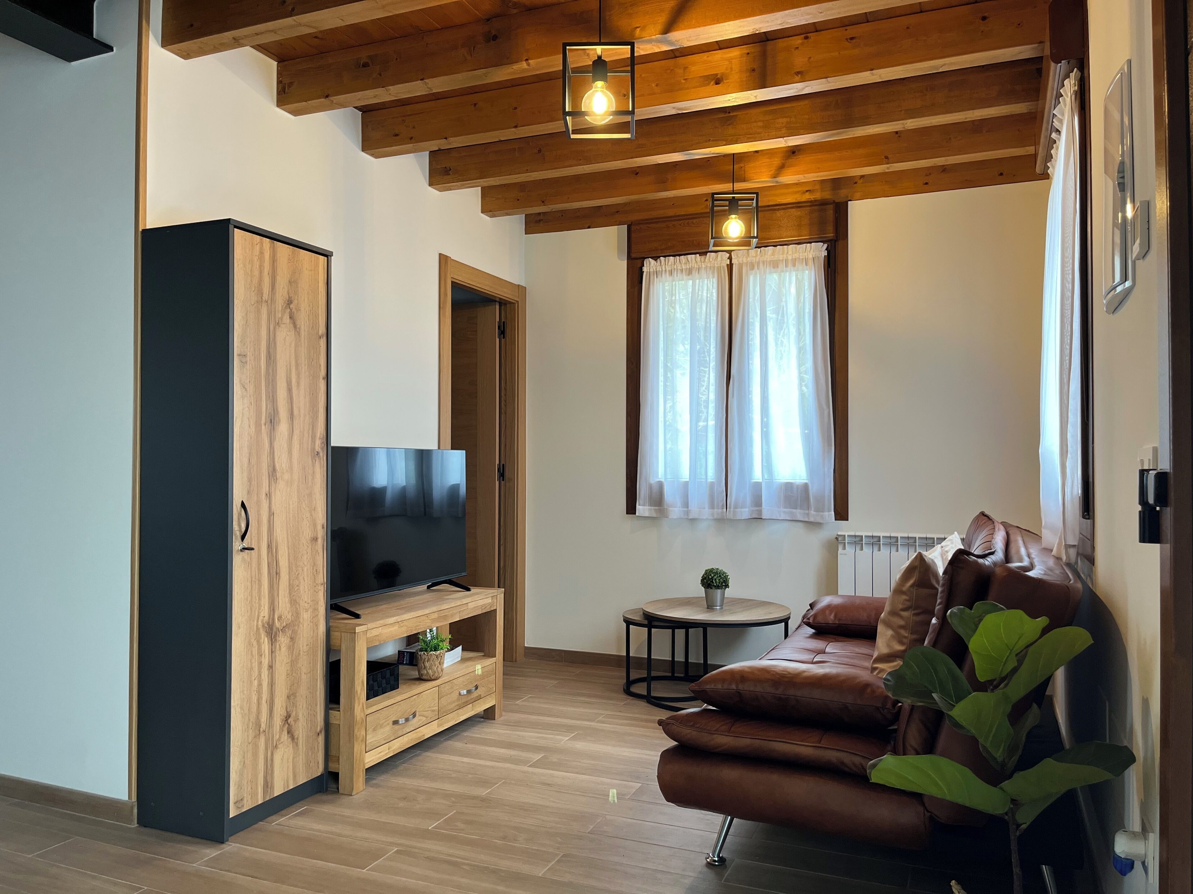 A modern living room with a wooden ceiling, a TV on a wooden stand, a leather sofa, a small round table, and a window with curtains.