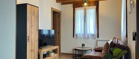 A modern living room with a wooden ceiling, a TV on a wooden stand, a leather sofa, a small round table, and a window with curtains.