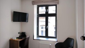 Quarto de hotel com cama, escrivaninha, cadeira, televisão na parede e janela com vista para prédios.