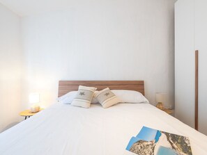 Ein ordentlich bezogenes Bett mit weißer Bettwäsche und gemusterten Kissen, ein hölzerner Kopfteil und ein kleiner Nachttisch mit einer Lampe.