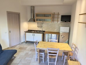 Eine kleine Küche mit Herd, Spüle und kleinem Kühlschrank. Ein Esstisch mit Stühlen und ein an der Wand befestigtes Fernsehgerät.