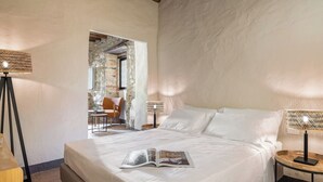 Ein Schlafzimmer mit einem großen Bett, zwei Nachttischlampen, einem kleinen Tisch mit Stuhl sowie Blick auf eine Steinsäule und eine Tür zu einem anderen Raum.