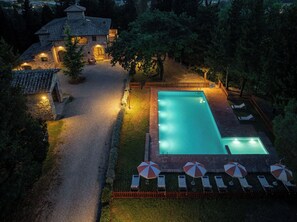 Piscina com espreguiçadeiras e guarda-sóis, cercada por um deck de tijolos e um edifício de pedra.