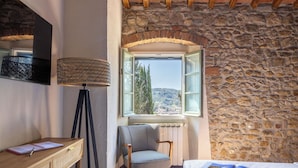 Ein Zimmer mit Steingemäuer, einem Fenster mit geöffnetem Rollo, einem Schreibtisch mit Lampe, einem Stuhl und einem Bett mit einer Zeitschrift.