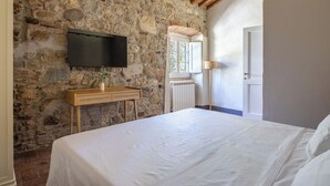 Un dormitorio con pared de piedra, techo de madera, cama, televisor, mesita de noche con un jarrón y ventana.