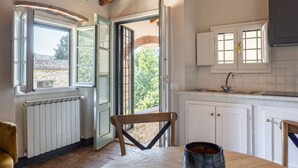 Una cocina con armarios blancos, una silla de madera, una mesa con un cuenco oscuro y vistas a través de puertas abiertas.