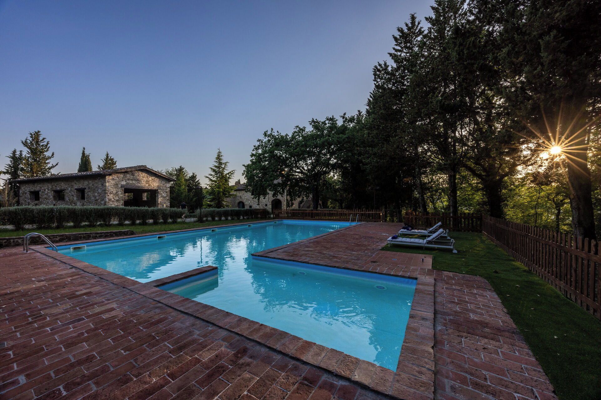 Una piscina rettangolare con bordo in mattoni, circondata da una recinzione di legno e alberi.