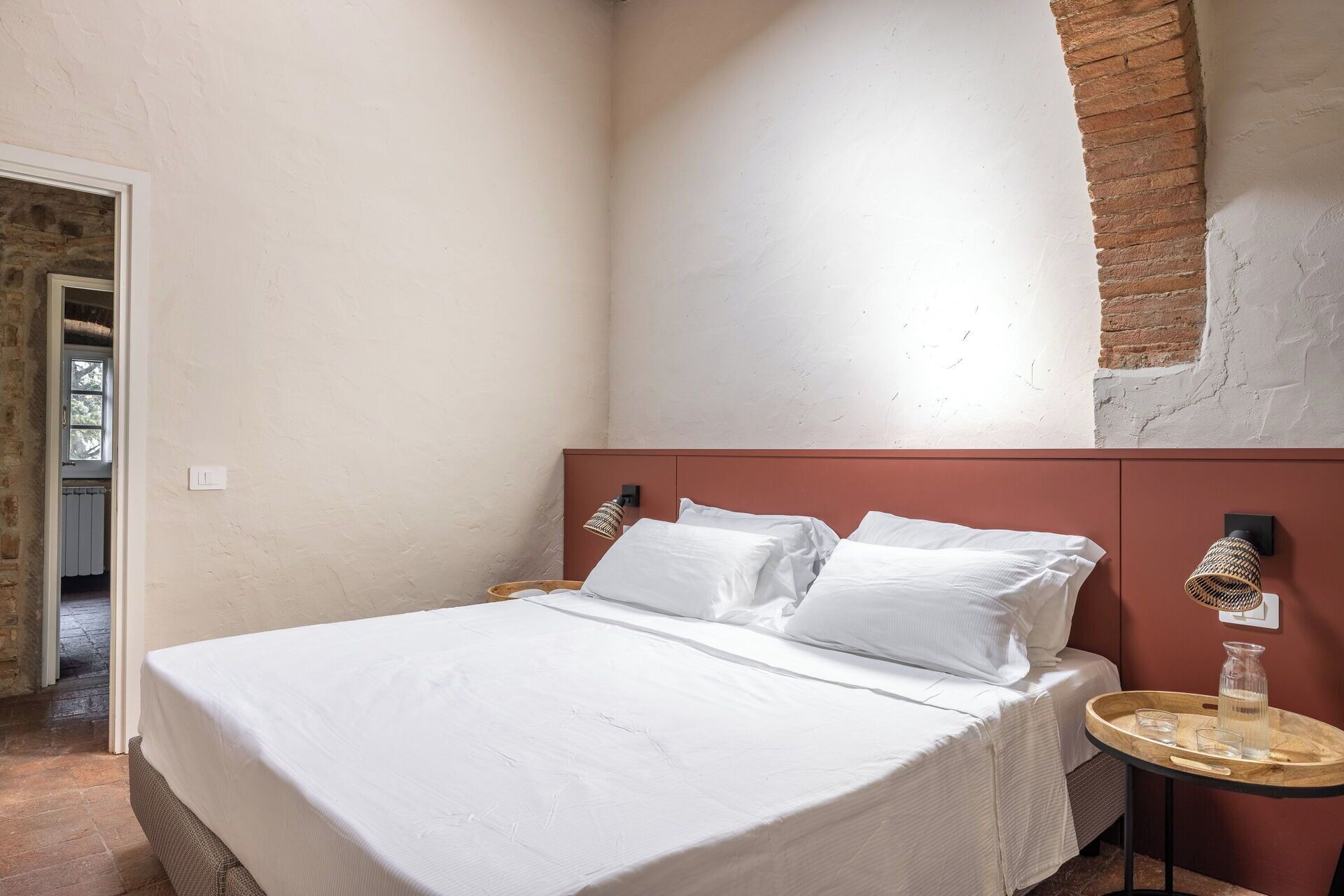 Un dormitorio con cama, mesita de noche, lámpara y vista a un muro de ladrillo.