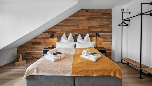 Ein modernes Schlafzimmer mit Schrägwand, einer Holzvertäfelung, einem Bett mit weißer und senfgelber Bettwäsche, einem Nachttisch, einer Bank und einer wandmontierten Leuchte.