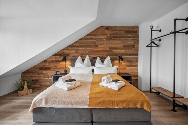 Ein modernes Schlafzimmer mit Schrägwand, einer Holzvertäfelung, einem Bett mit weißer und senfgelber Bettwäsche, einem Nachttisch, einer Bank und einer wandmontierten Leuchte.