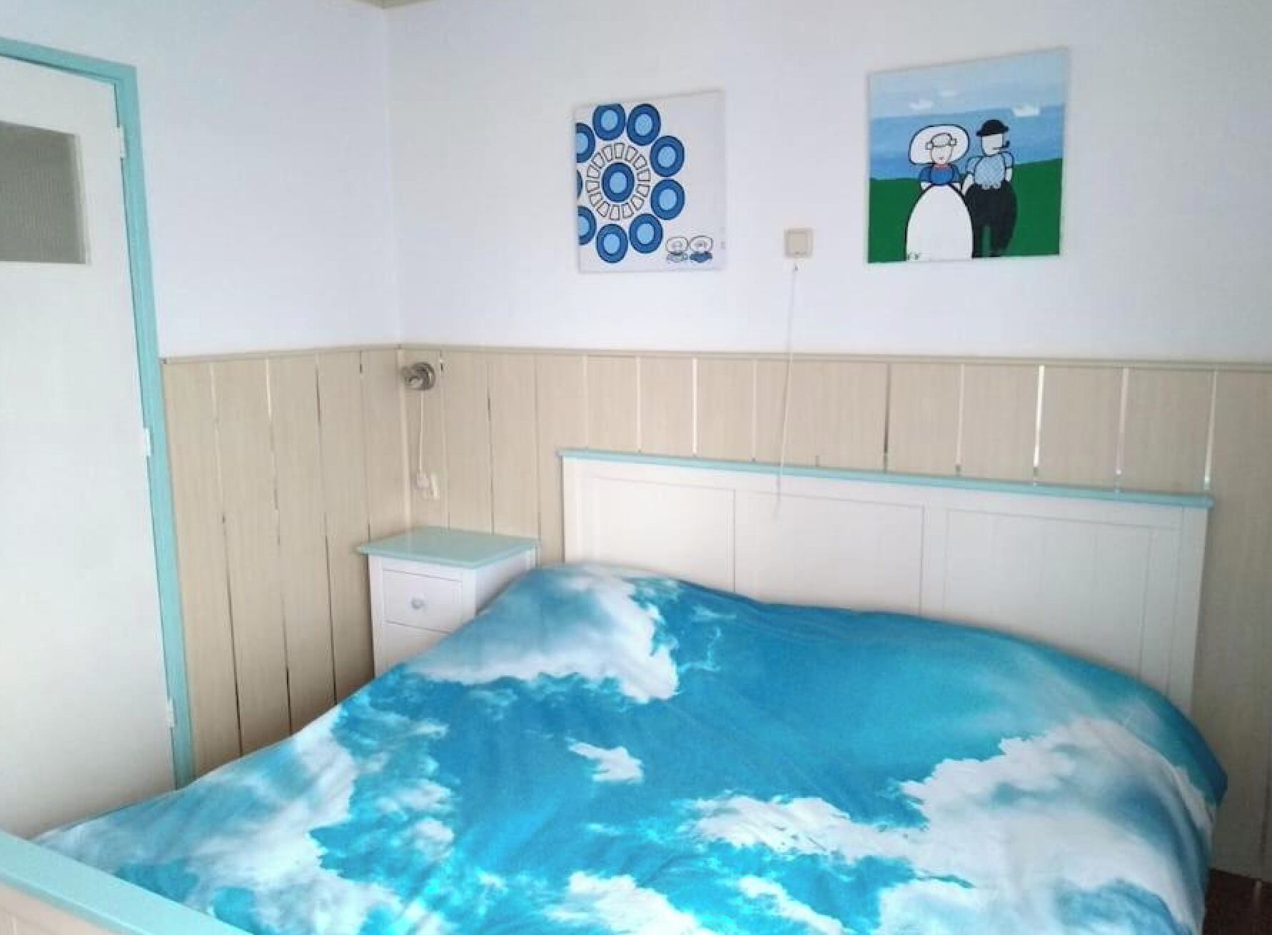 Een slaapkamer met een bed met een motief van lucht en wolken, een nachtkastje, een in de muur gemonteerde lichtschakelaar en twee ingelijste kunstwerken.