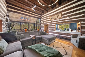 Ein Holzblockhaus-Innenraum mit einem Ecksofa, einem grünen Pouf und einer Küchenzeile mit blauen Schränken.