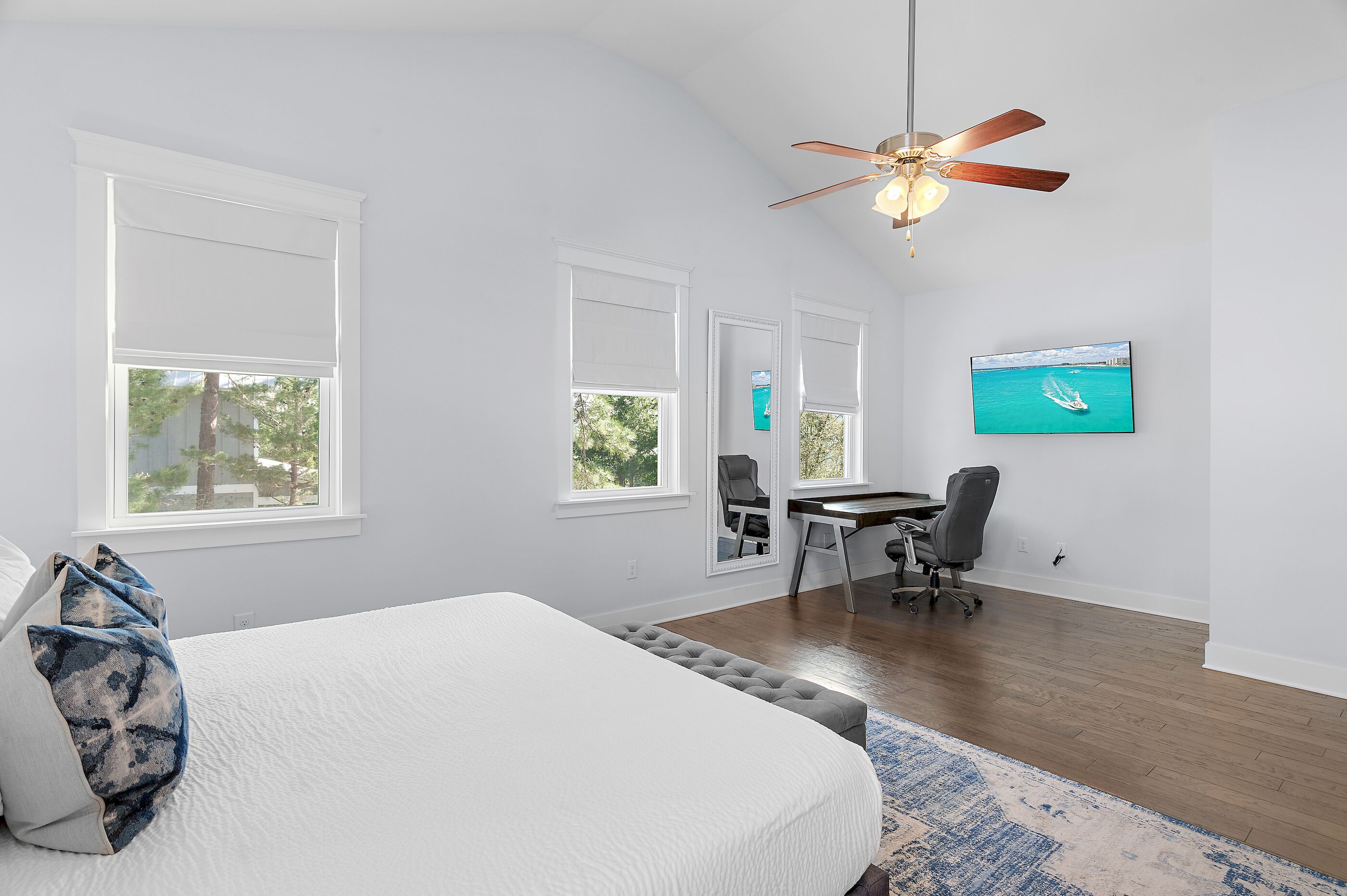 A bedroom with a bed, a desk, a chair, a ceiling fan, and a TV on the wall.