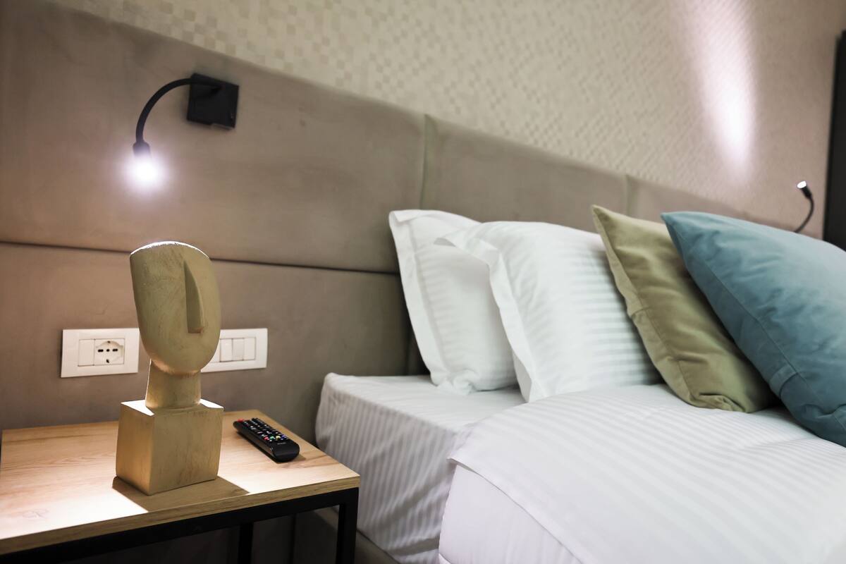 A hotel room with a bed, a bedside table with a remote control, a wall-mounted lamp, and an outlet.