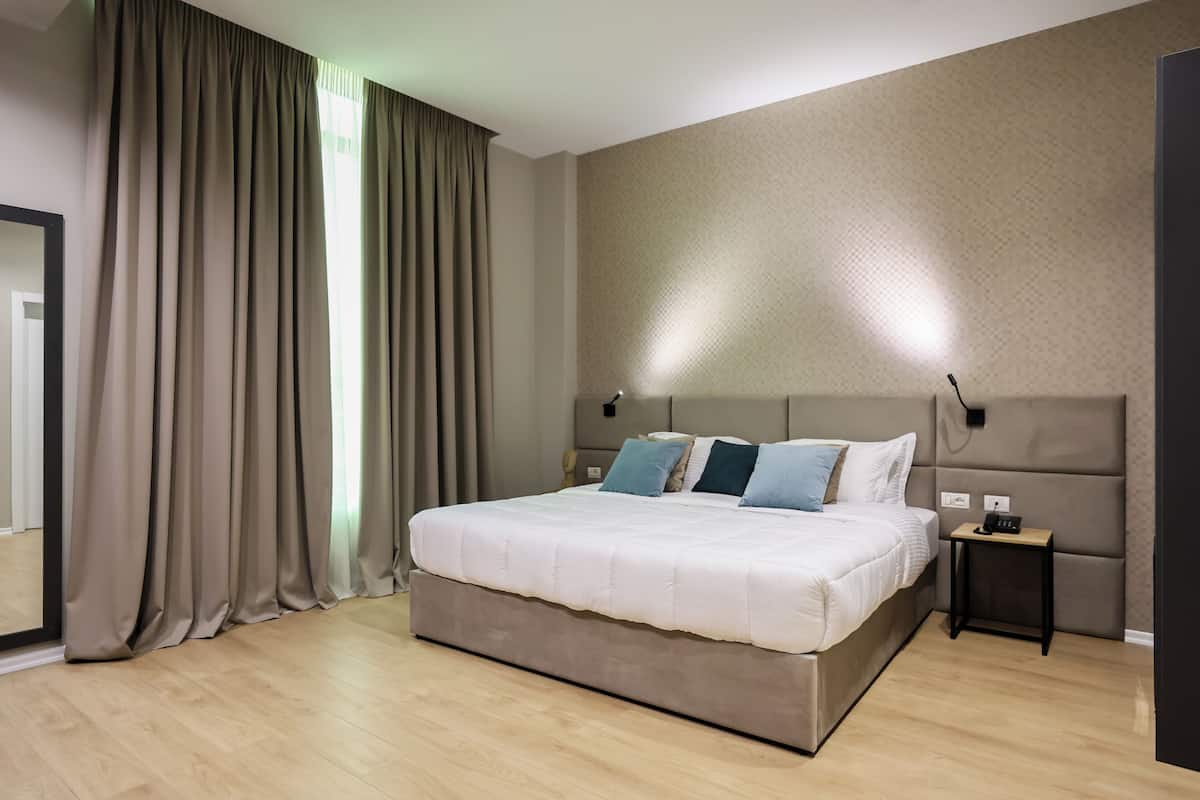 A modern hotel room with a large bed, a nightstand, a wall-mounted lamp, and a large mirror.