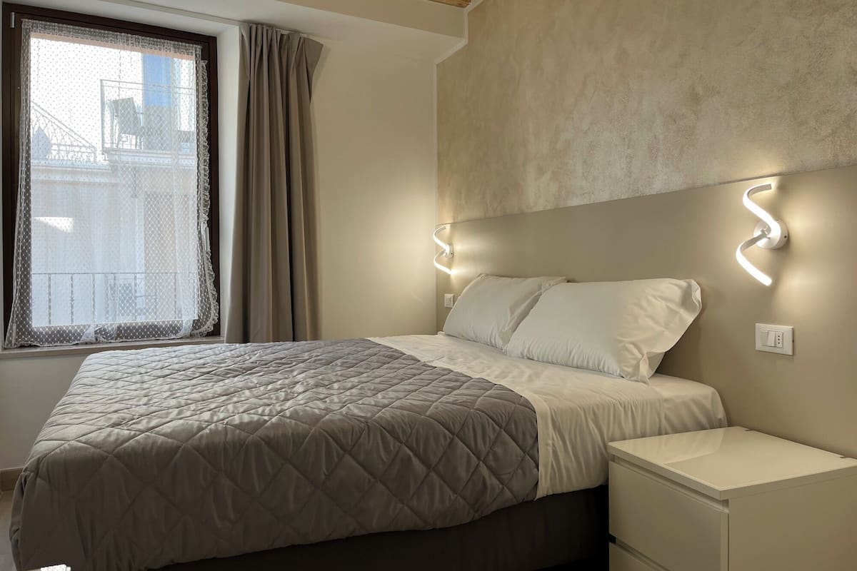 Ein Schlafzimmer mit Bett, Nachttischen, einem Fenster mit Vorhängen und Wandleuchten.