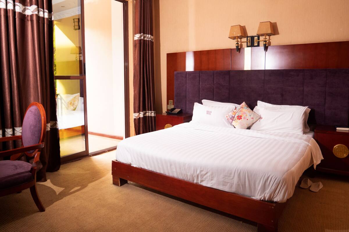 Una habitación de hotel con una cama grande, cabecero de madera, mesita de noche, una silla y puerta corredera que da a un balcón.