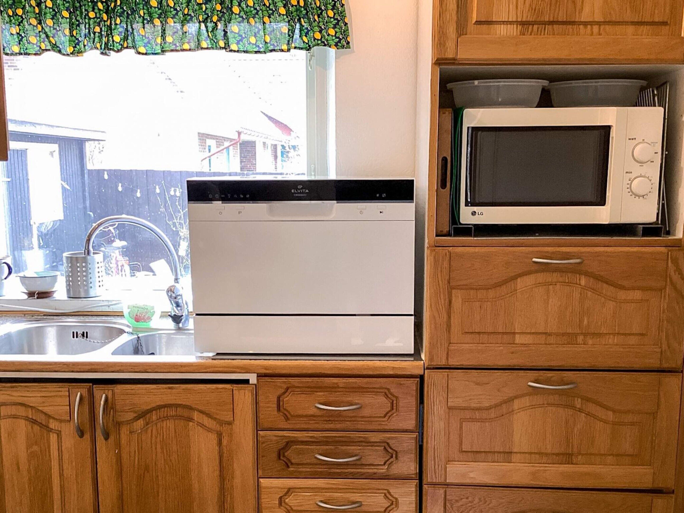 Une cuisine équipée d’un lave-vaisselle, d’un four à micro-ondes, de meubles en bois et d  ’une fenêtre agrémentée d’un rideau à motifs.