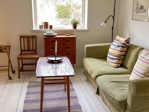 Ein Zimmer mit einem Holztisch, einem grünen Sofa, einem Sessel, einem Schminktisch und einem Fenster mit Blick auf Bäume.