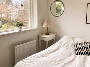 Ein Schlafzimmer mit einem Bett, einem Nachttisch mit Lampe, einem Fenster mit Blick ins Grüne und einem Wandspiegel.