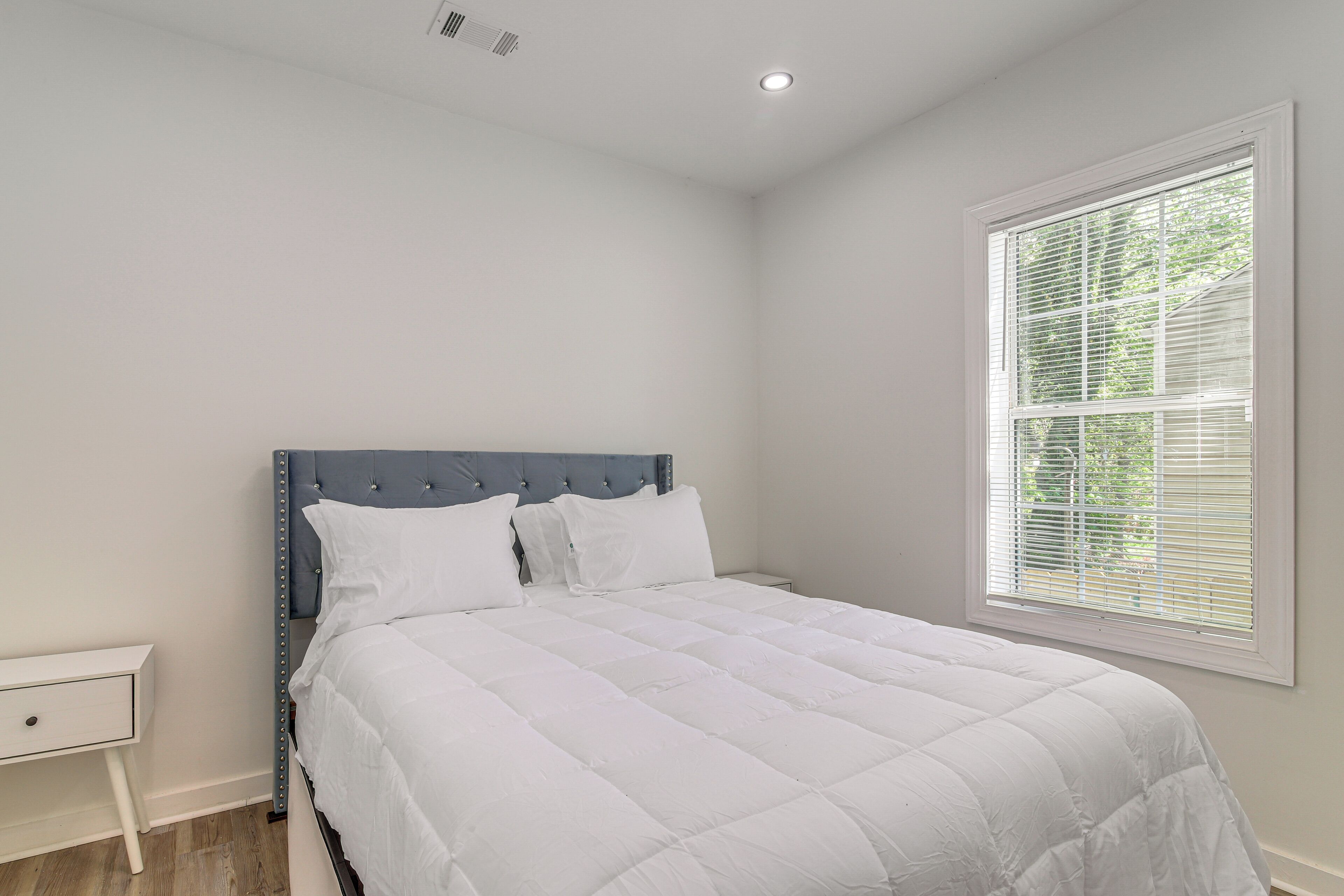Une chambre à coucher comprenant un lit, une table de chevet, une fenêtre avec des stores et une bouche d’aération au plafond.