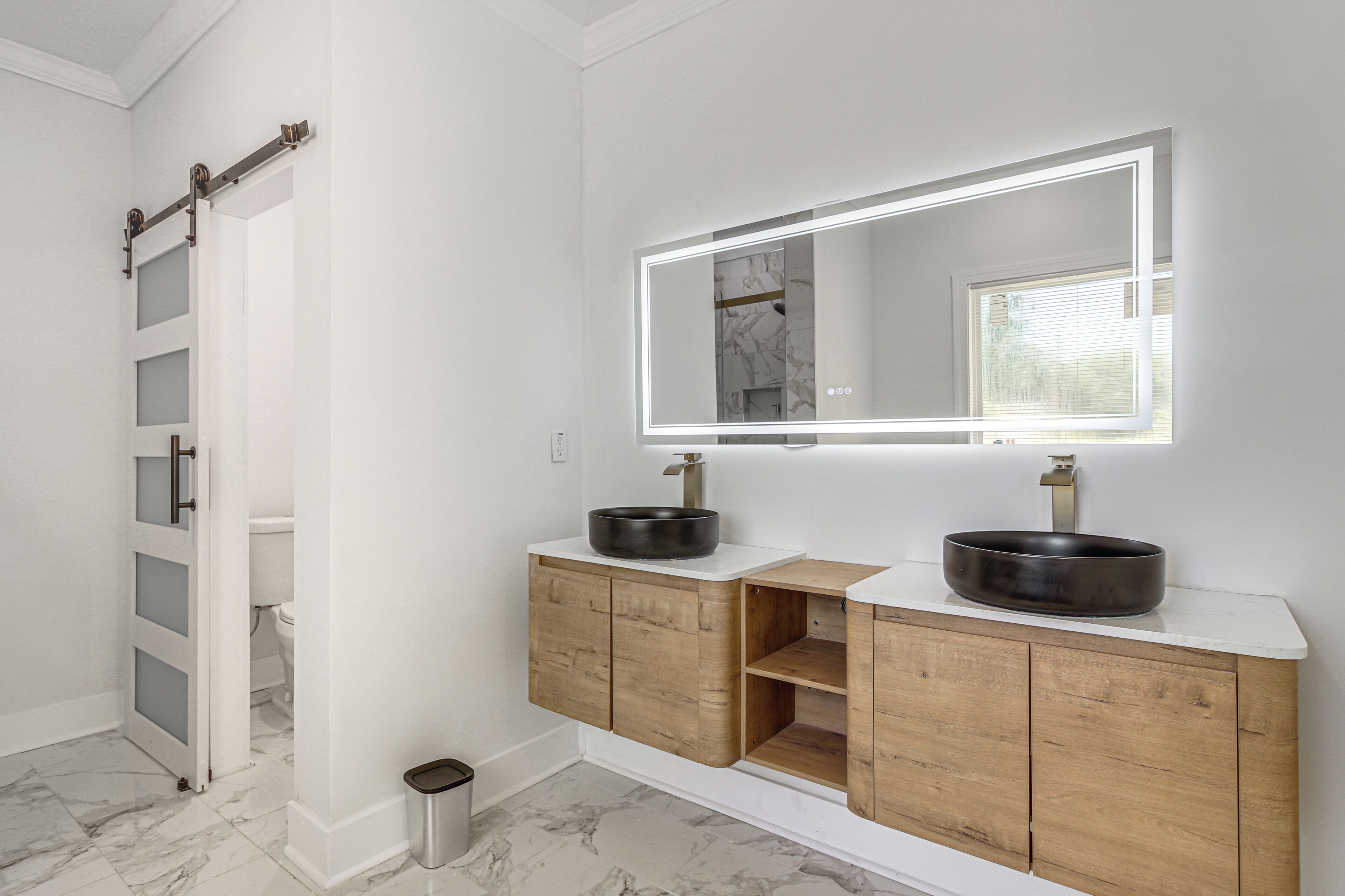 A bathroom with a white vanity, two black sinks, a mirror with lighting, wooden cabinets, and a marble floor.
