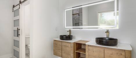 A bathroom with a white vanity, two black sinks, a mirror with lighting, wooden cabinets, and a marble floor.