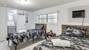 A bedroom with two beds, a window, a ceiling fan, a wall-mounted air conditioner, and a small bedside table with a plant.