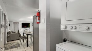 A laundry room with a Whirlpool washer and dryer, a fire extinguisher, a dining area with a table and chairs, and a living area with a sofa and a television.