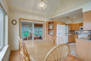 Uma sala de jantar com mesa e cadeiras de madeira, uma cozinha com armários de madeira, uma geladeira e um relógio na parede.