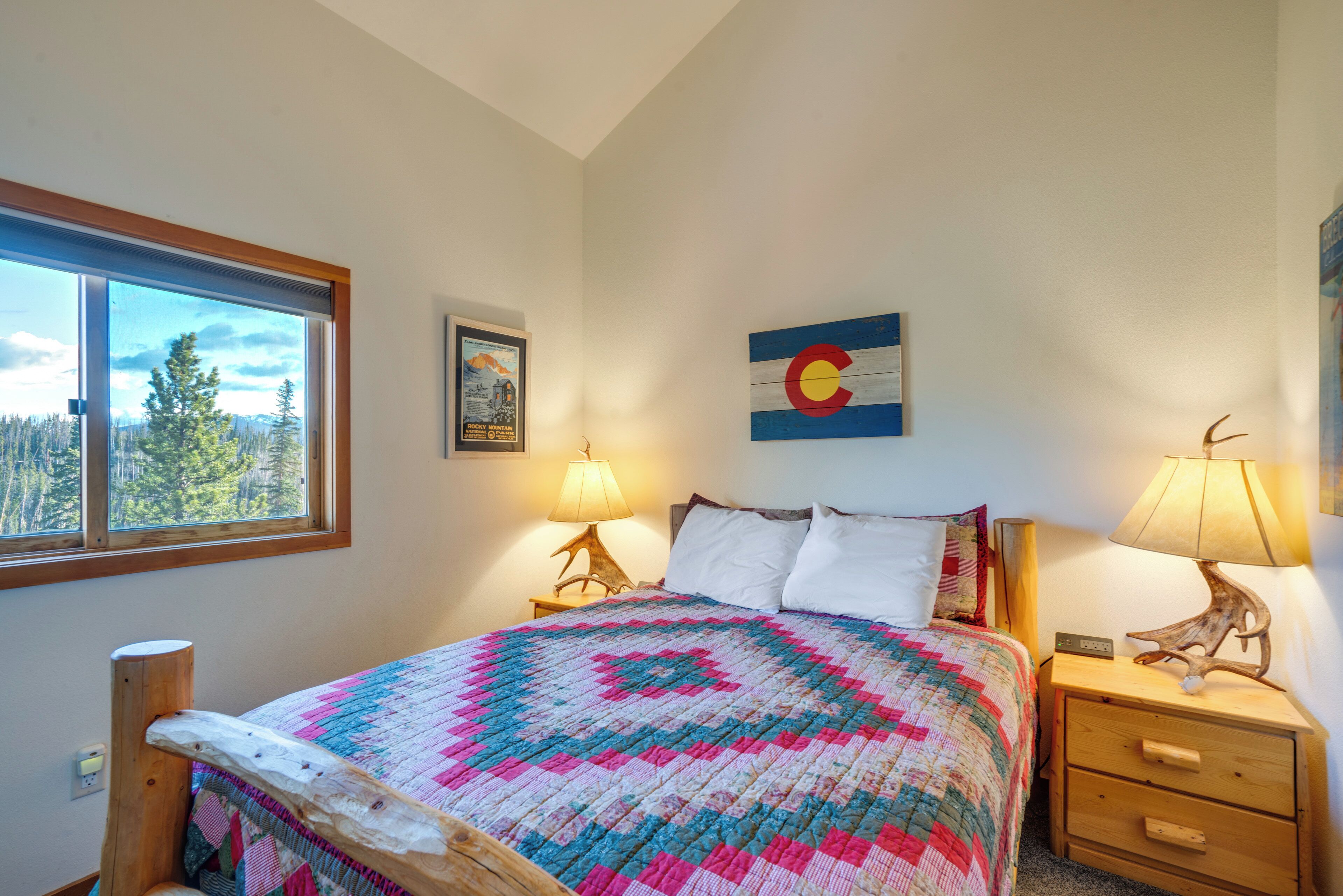 Une chambre avec une grande fenêtre, un lit recouvert d’une couverture aux couleurs vives, une table de chevet en bois, une lampe et un tableau accroché au mur.