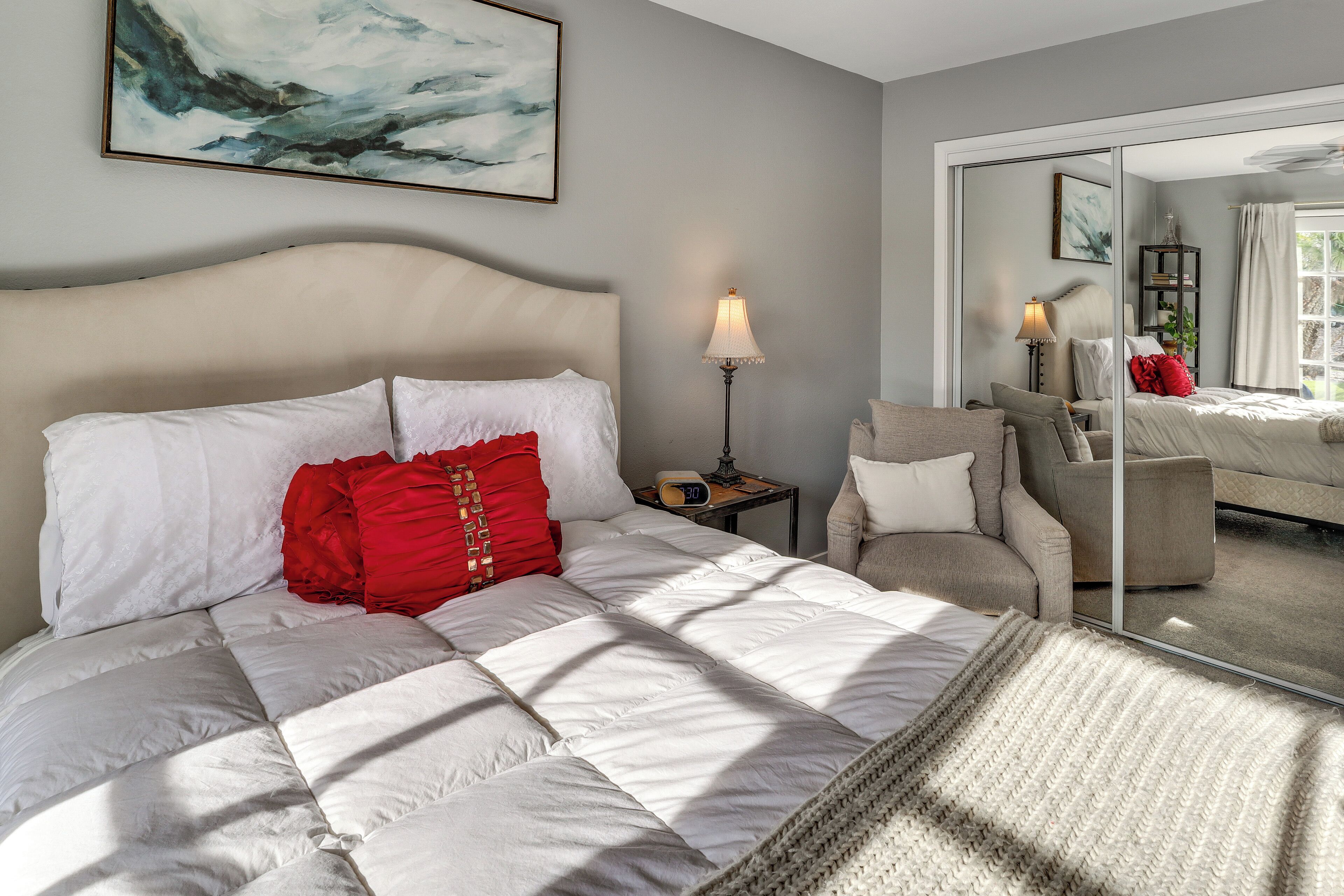 Ein Schlafzimmer mit Bett, Nachttisch, Lampe, Stuhl, Spiegel und einem Bild an der Wand.