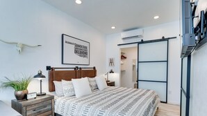 Un dormitorio con cama, mesita de noche, lámpara, una obra de arte en la pared y una planta.