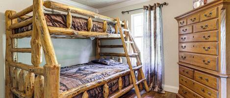 A bunk bed with a ladder, a wooden dresser, and a ceiling fan in a room with wooden flooring and a window with curtains.