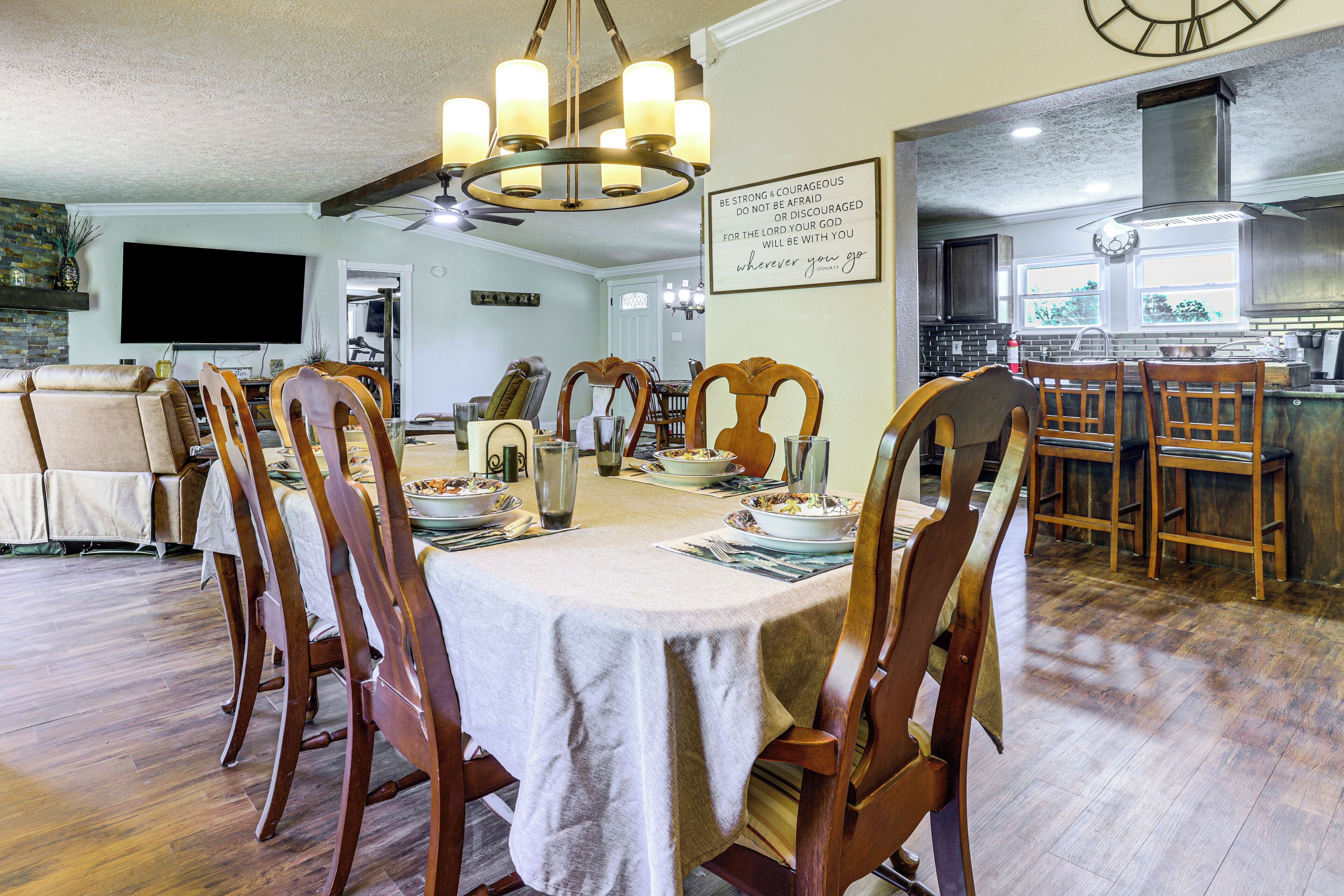 Un coin repas avec une table dressée pour six personnes, des chaises en bois, un lustre et une cuisine en arrière-plan.