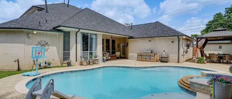 A residential swimming pool with a covered patio area, a basketball hoop, and a brick exterior.