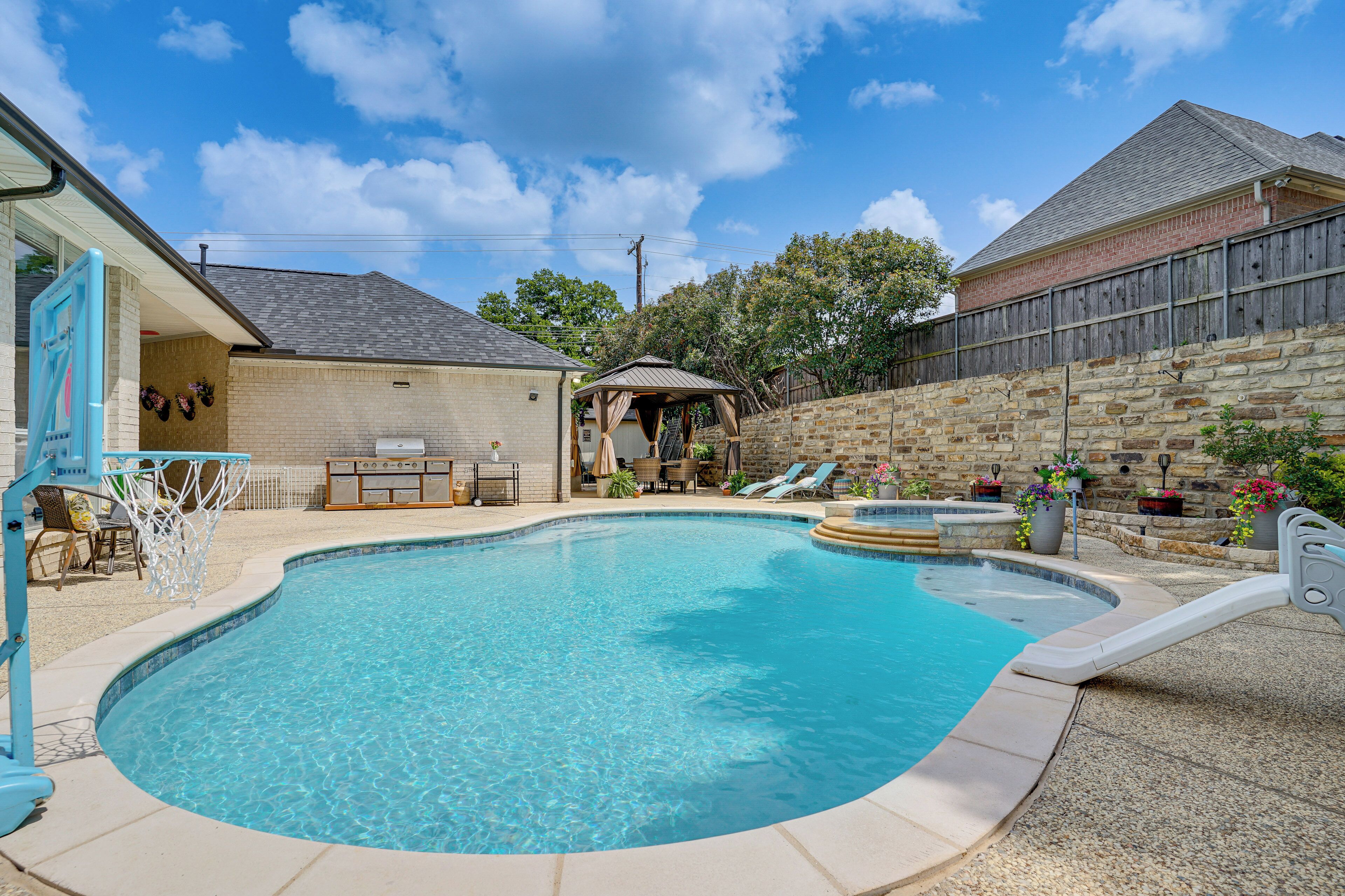 A backyard with a swimming pool, a covered patio, and a stone wall.