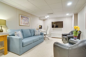 Ein Wohnzimmer mit einem blauen Sofa, einem schwarzen Ledersessel, einem Holztischchen, einem weißen Schreibtisch und einem an der Wand befestigten Fernseher.