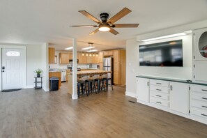 A modern kitchen with wooden cabinets, a central island with bar stools, a ceiling fan, and a large TV mounted on the wall.